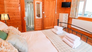 A wooden cabin bedroom with a bed, a TV, a shower, and a window with curtains. - Borah Lodge (Penzance)