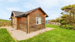 A wooden cabin with a stone wall, a small porch, and a grassy yard. - Borah Lodge (Penzance)