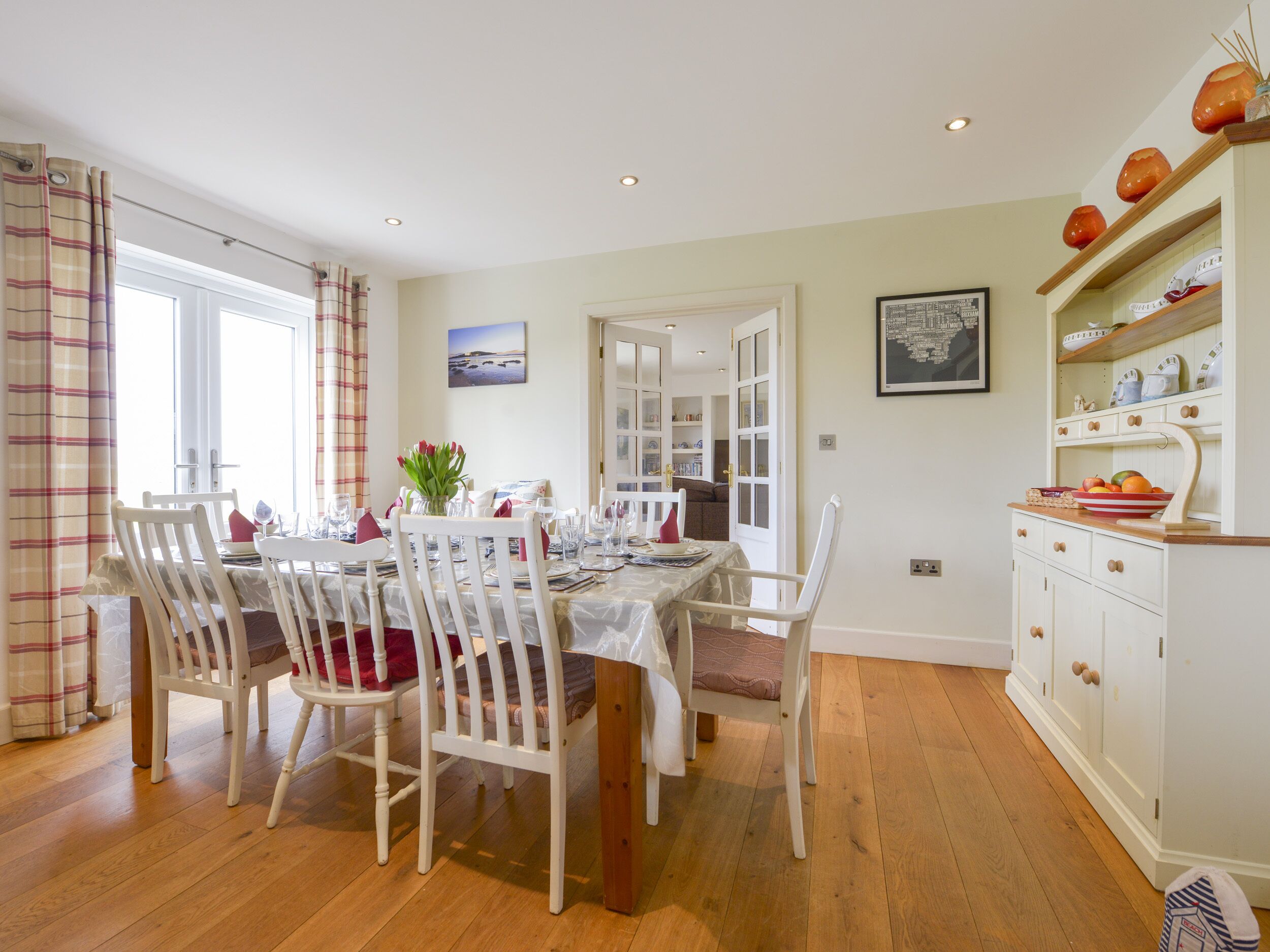 A dining area with a table set for four, surrounded by white chairs with red cushions. There is a window with curtains, a painting on the wall, and a kitchen area with cabinets and a shelf.