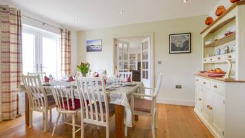 A dining area with a table set for four, surrounded by white chairs with red cushions. There is a window with curtains, a painting on the wall, and a kitchen area with cabinets and a shelf.