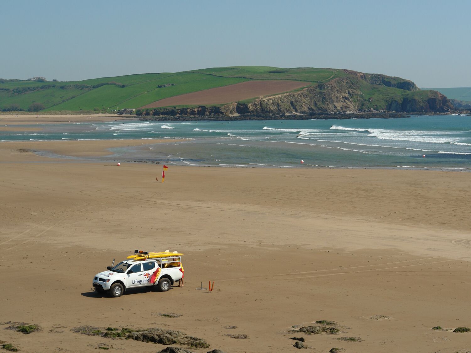 A lifeguard vehicle parked on a sandy beach with a rocky cliff and ocean in the background.
