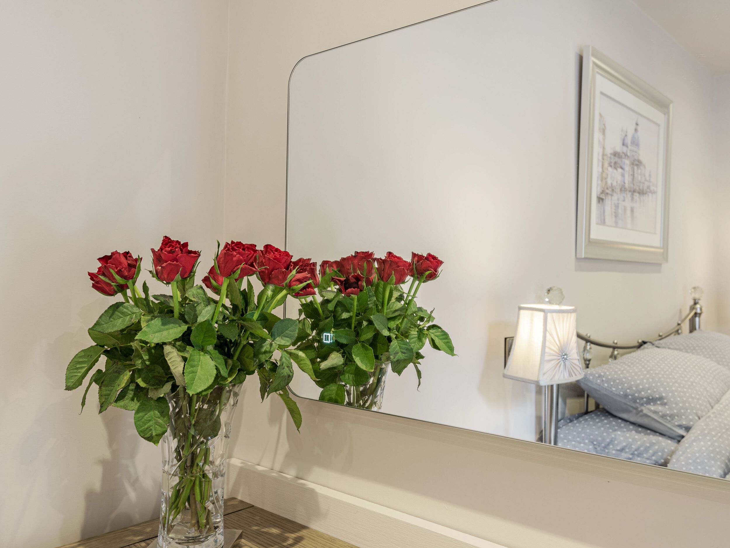 A bathroom with a mirror, a vase of red roses, a bed with grey bedding, and a framed picture on the wall.