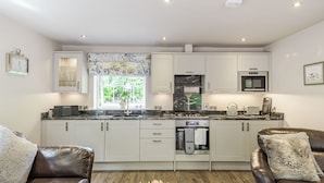 A modern kitchen with white cabinets, a built-in oven, and a microwave. There is a window with a view of greenery outside. - Silvadale Cottage (Louth)