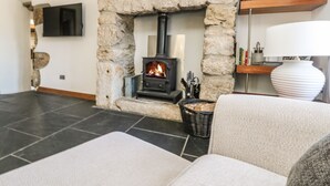 A living room with a stone fireplace, a flat-screen TV mounted on the wall, and a bookshelf with books and decorative items. - Miner's Rest (Camborne)