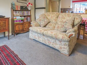 A cozy living room with a floral sofa, a wooden bookshelf, and a kitchen area visible through an open doorway. - River Cottage (Tewkesbury)