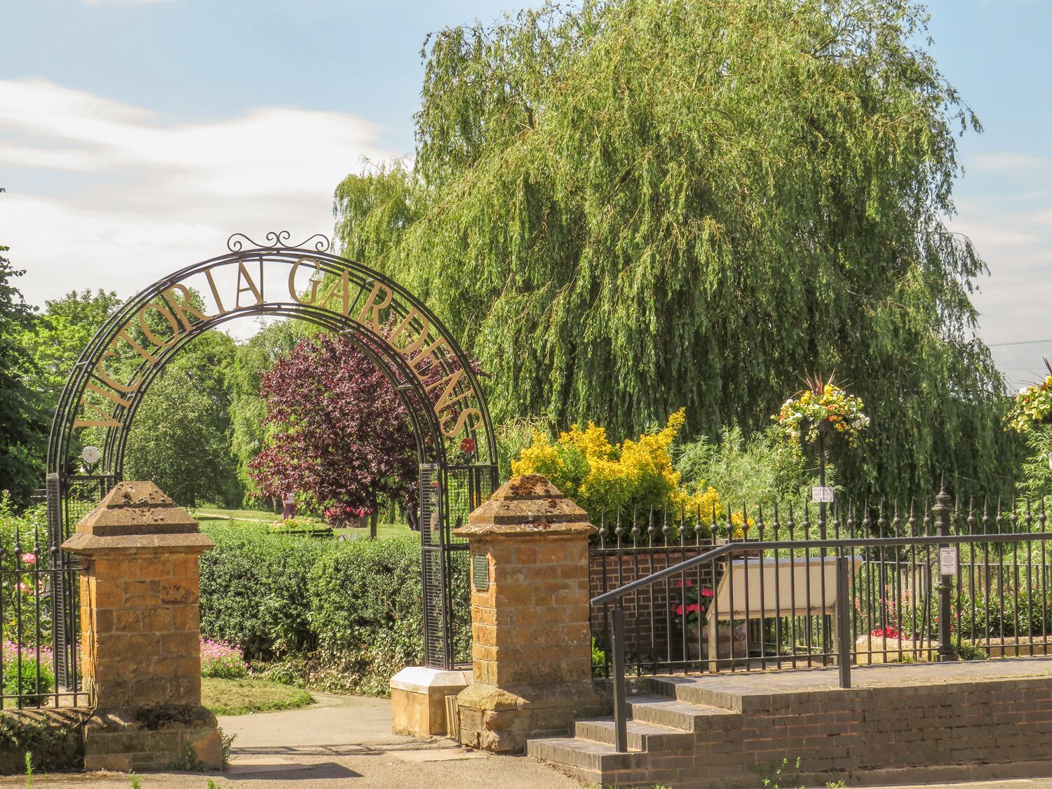 A wrought iron gate with the inscription "GARDENS" arching over a pathway leading into a park.