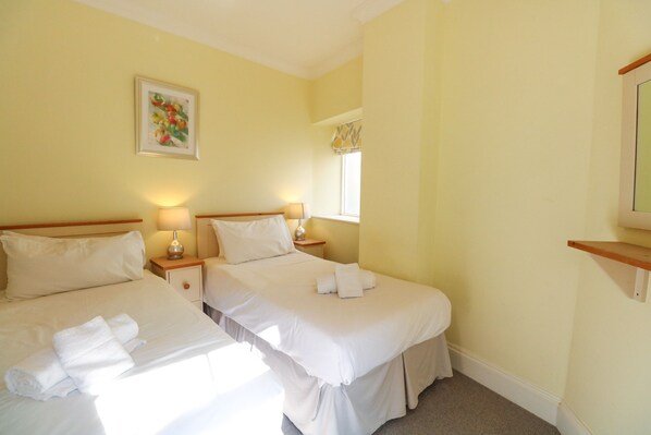 A hotel room with two beds, a bedside table with a lamp, a window with a curtain, and a wall-mounted shelf.