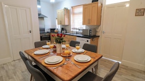 A dining area with a wooden table set for four, surrounded by chairs. There is a kitchen in the background with wooden cabinets and a microwave on the counter. - The Tannery (Whitby)