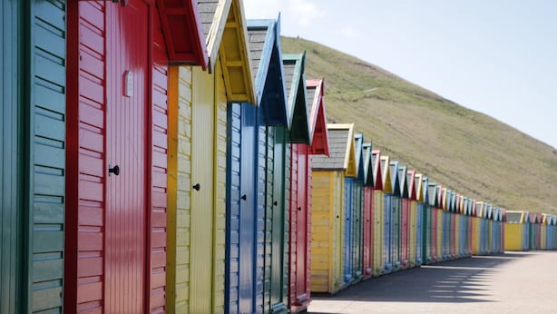 A row of colorful beach huts with sloped roofs, arranged along a paved path beside a hill. - The Tannery (Whitby)