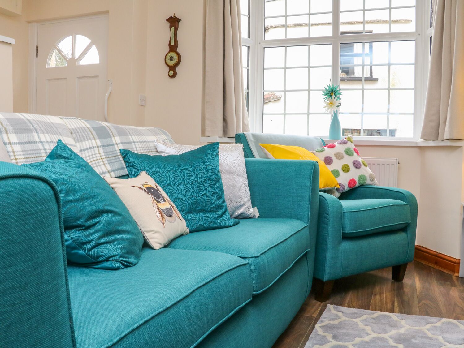 A teal sofa with patterned cushions, a window with a view, and a wall clock.