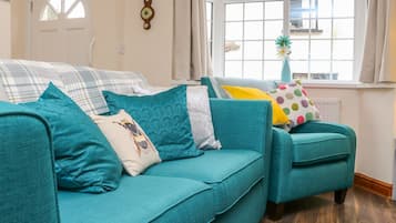 A teal sofa with patterned cushions, a window with a view, and a wall clock.
