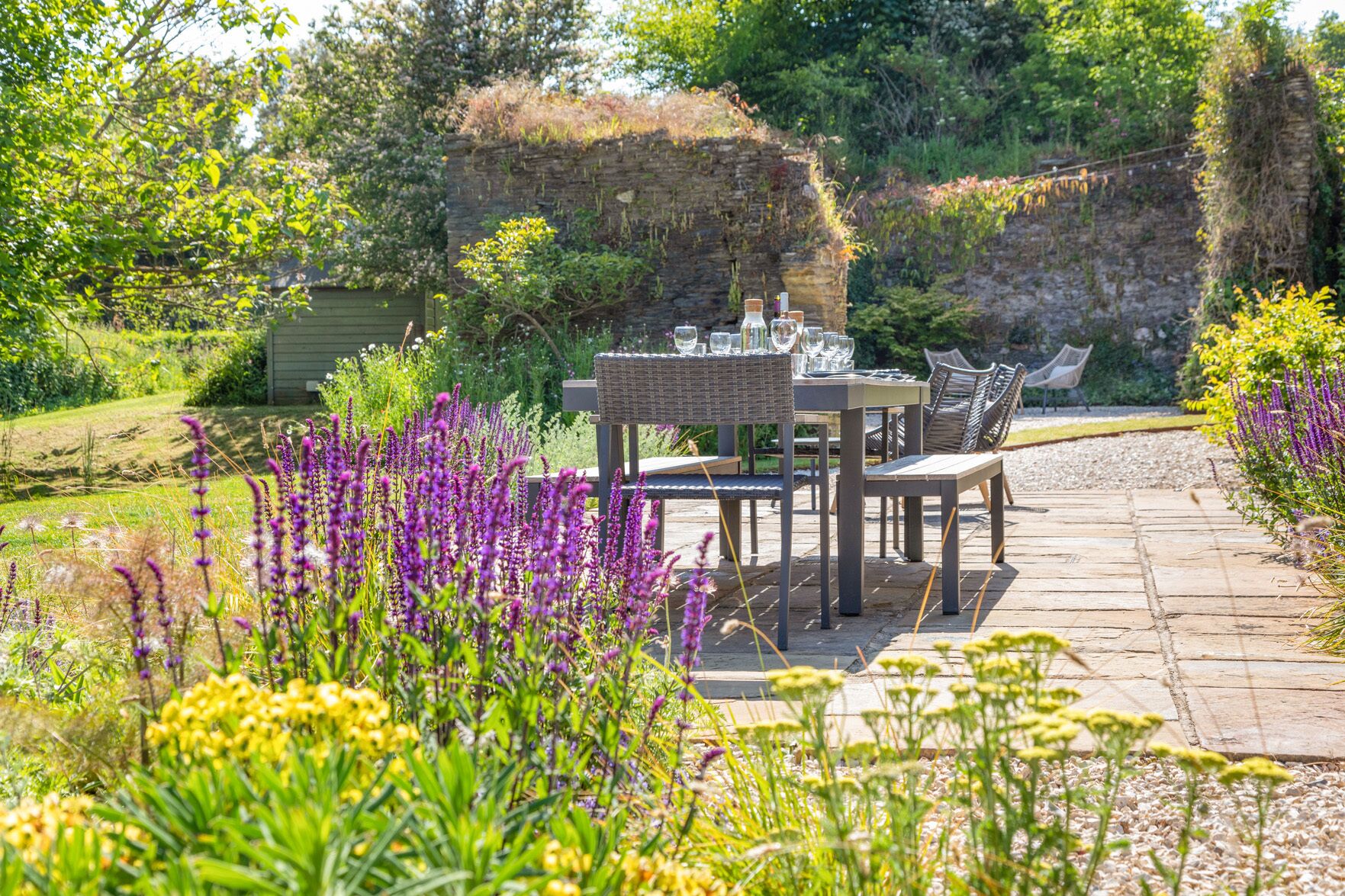 An outdoor dining area with a stone table and chairs, surrounded by lush greenery and flowering plants.