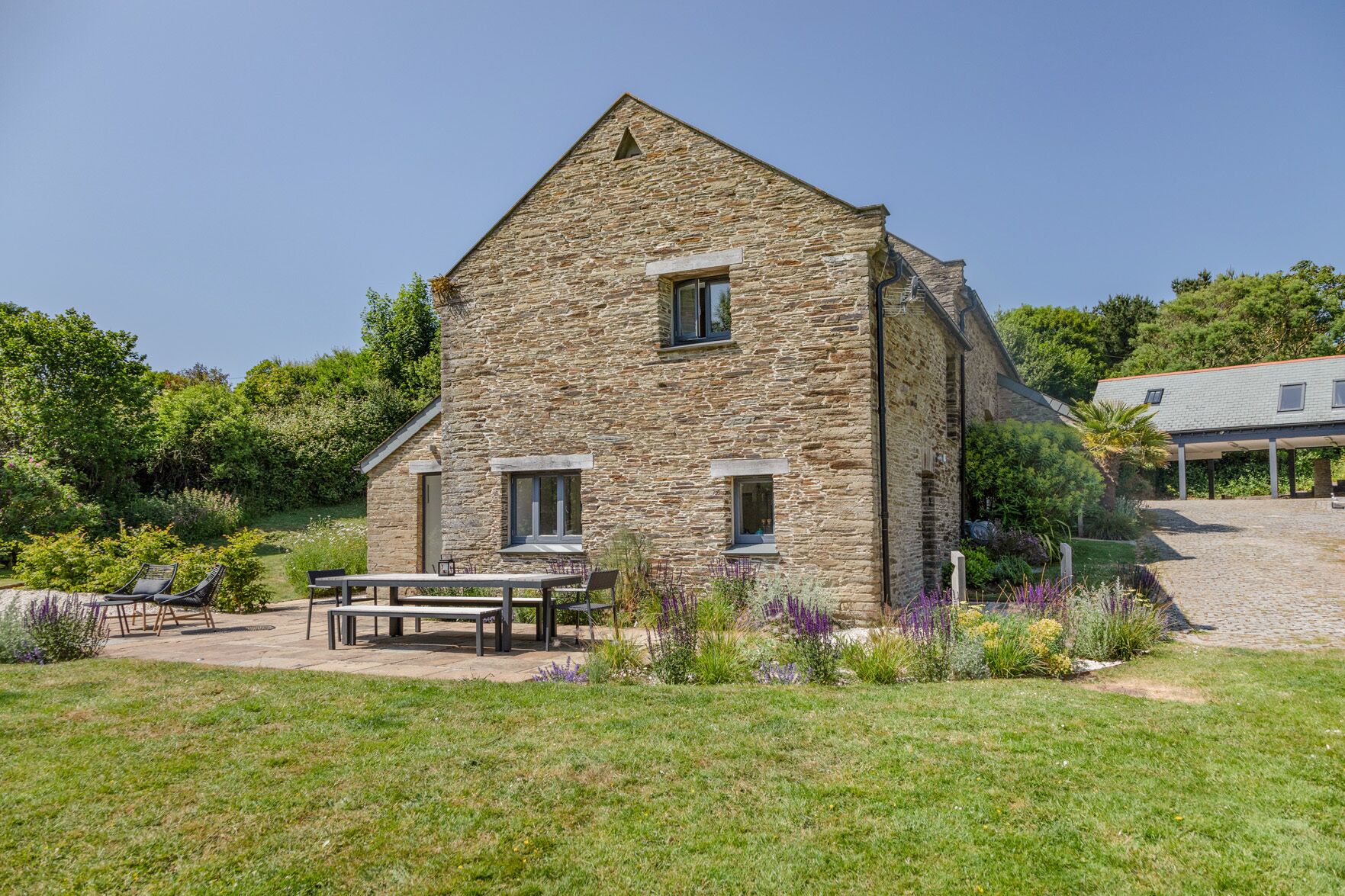 A stone building with a sloped roof, a covered patio with outdoor seating, and a garden with various plants and flowers.