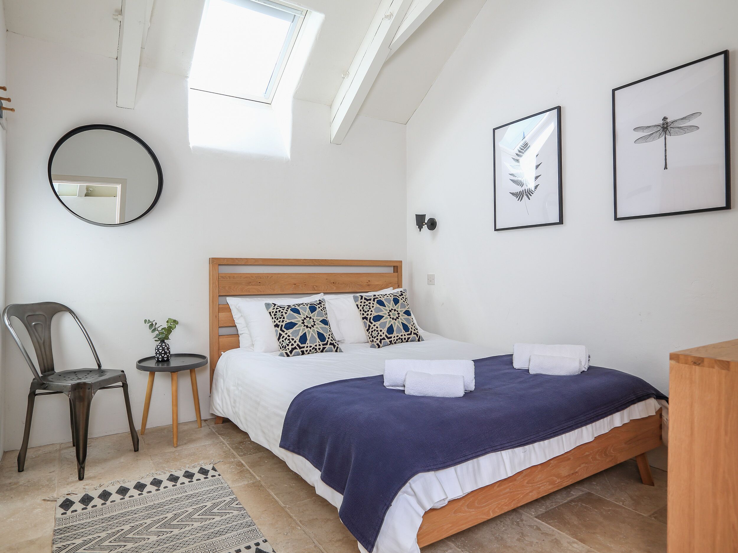 Une chambre avec un lit en bois, un miroir rond, une petite table avec une plante et une chaise. Deux tableaux encadrés sont accrochés au mur et une lucarne se trouve au-dessus du lit.