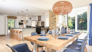 Una cocina moderna con una isla central, un comedor con mesa y sillas de madera, y un gran ventanal con vistas a zonas verdes.