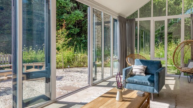 Una habitación moderna con grandes puertas de vidrio que dan a un jardín, un sofá azul, una silla de mimbre y una mesa de centro de madera.