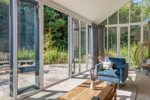 A modern room with large glass doors leading to a garden, a blue sofa, a wicker chair, and a wooden coffee table. - Linham Barn (Kingsbridge)