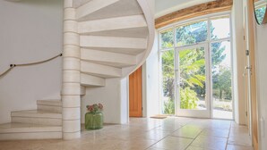 Una habitación espaciosa con una escalera de caracol, un amplio ventanal que da a un espacio exterior y un jarrón verde con flores sobre un pedestal.