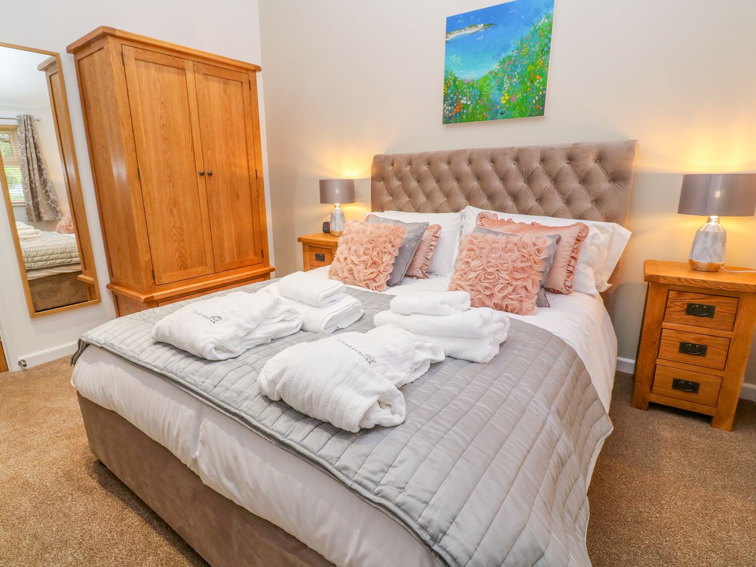 A neatly made bed with a tufted headboard, two bedside tables with lamps, a wooden wardrobe, and a painting on the wall.