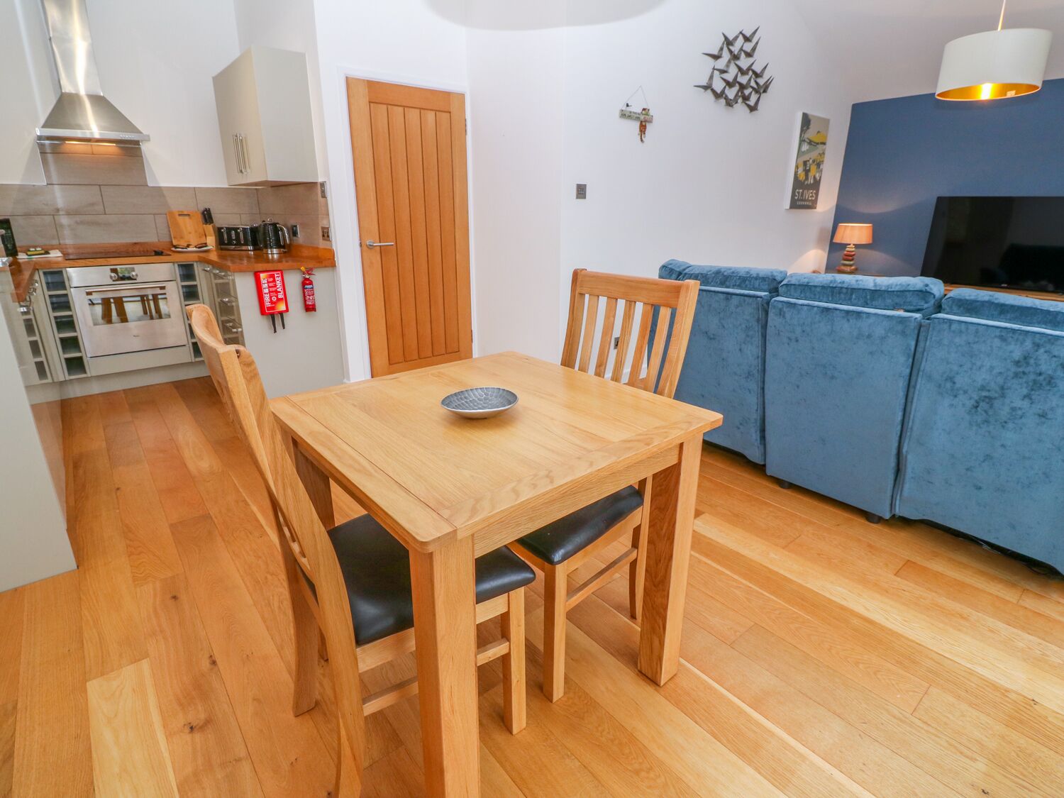A compact living space with a wooden dining table and chairs, a kitchenette, a blue sofa, and a TV.