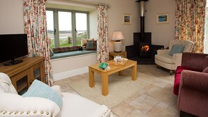 Una sala de estar acogedora con un sofá, un sillón y una mesita con una lámpara. Hay un televisor sobre un mueble bajo, una chimenea y una ventana con cortinas.