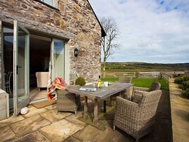 Una casa de piedra con una mesa de patio y sillas de mimbre, una puerta de cristal que da al interior, y vistas al campo.