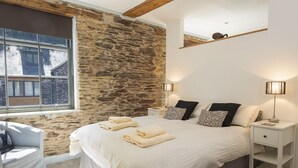 A bedroom with a stone wall, a large bed with white bedding, two bedside tables with lamps, and a window with a view of buildings.