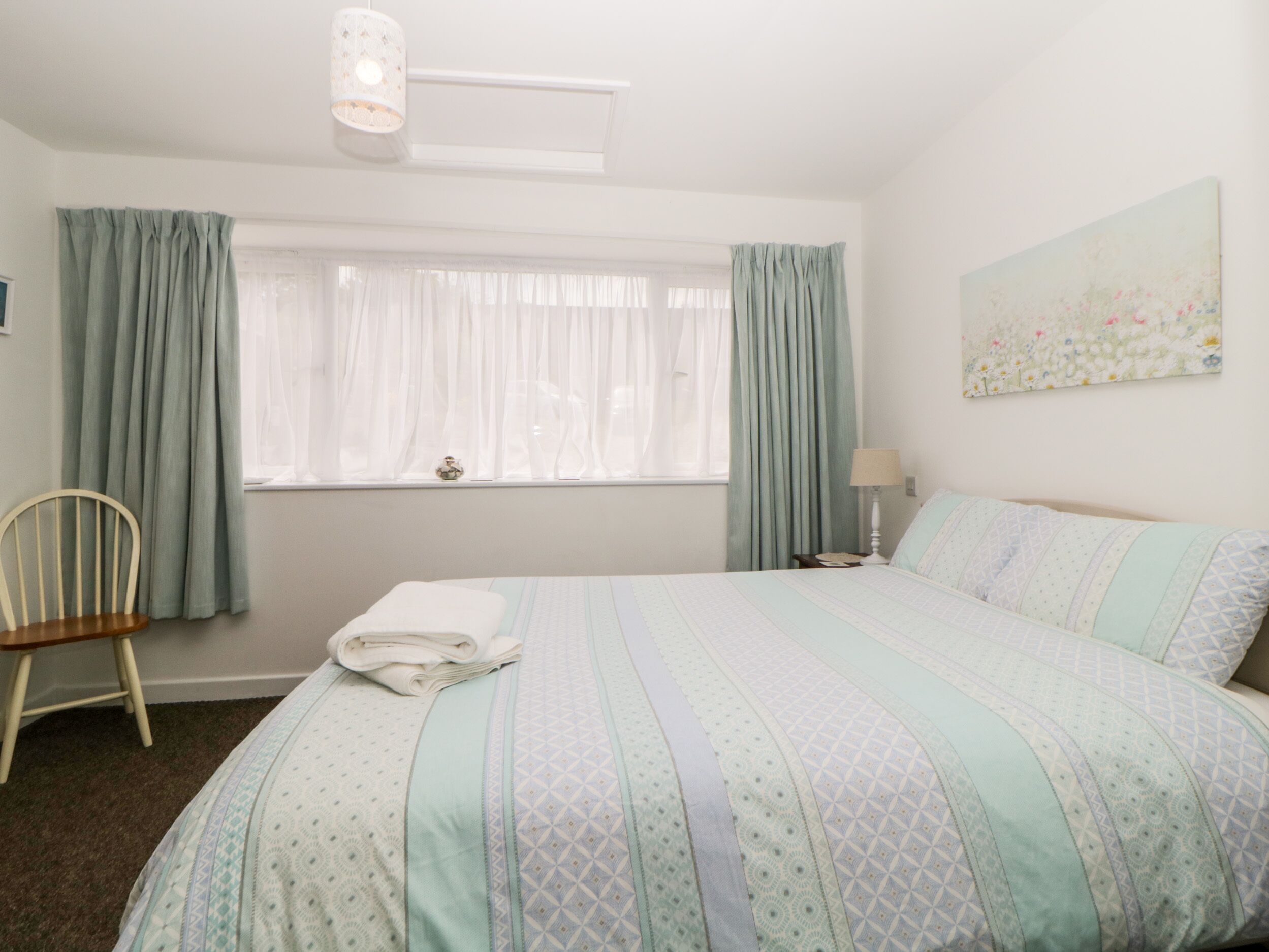 A neatly arranged bedroom with a bed, a chair, a window with curtains, a bedside table with a lamp, and a wall-mounted painting.