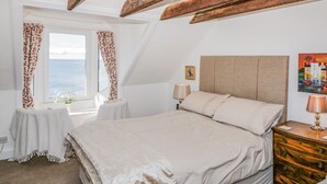 A bedroom with a large bed, wooden beams, a window with curtains, a bedside table with a lamp, and a painting on the wall.