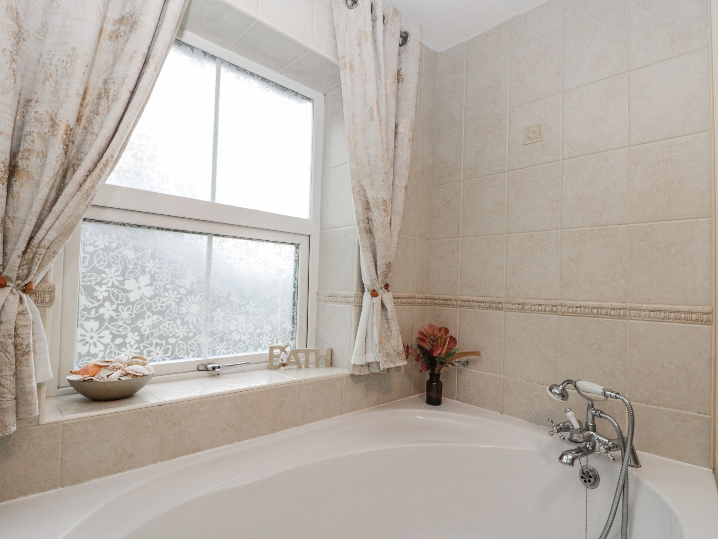 A bathroom with a bathtub, a window with curtains, and a vase with flowers on the windowsill.