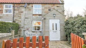 A stone house with a red-tiled roof, a white door, and a small garden area with a wooden fence and metal chairs. - Dove Cottage (Whitby)