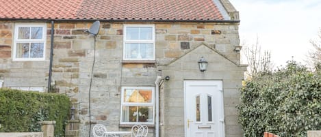 A stone house with a red-tiled roof, a white door, and a small garden area with a wooden fence and metal chairs.