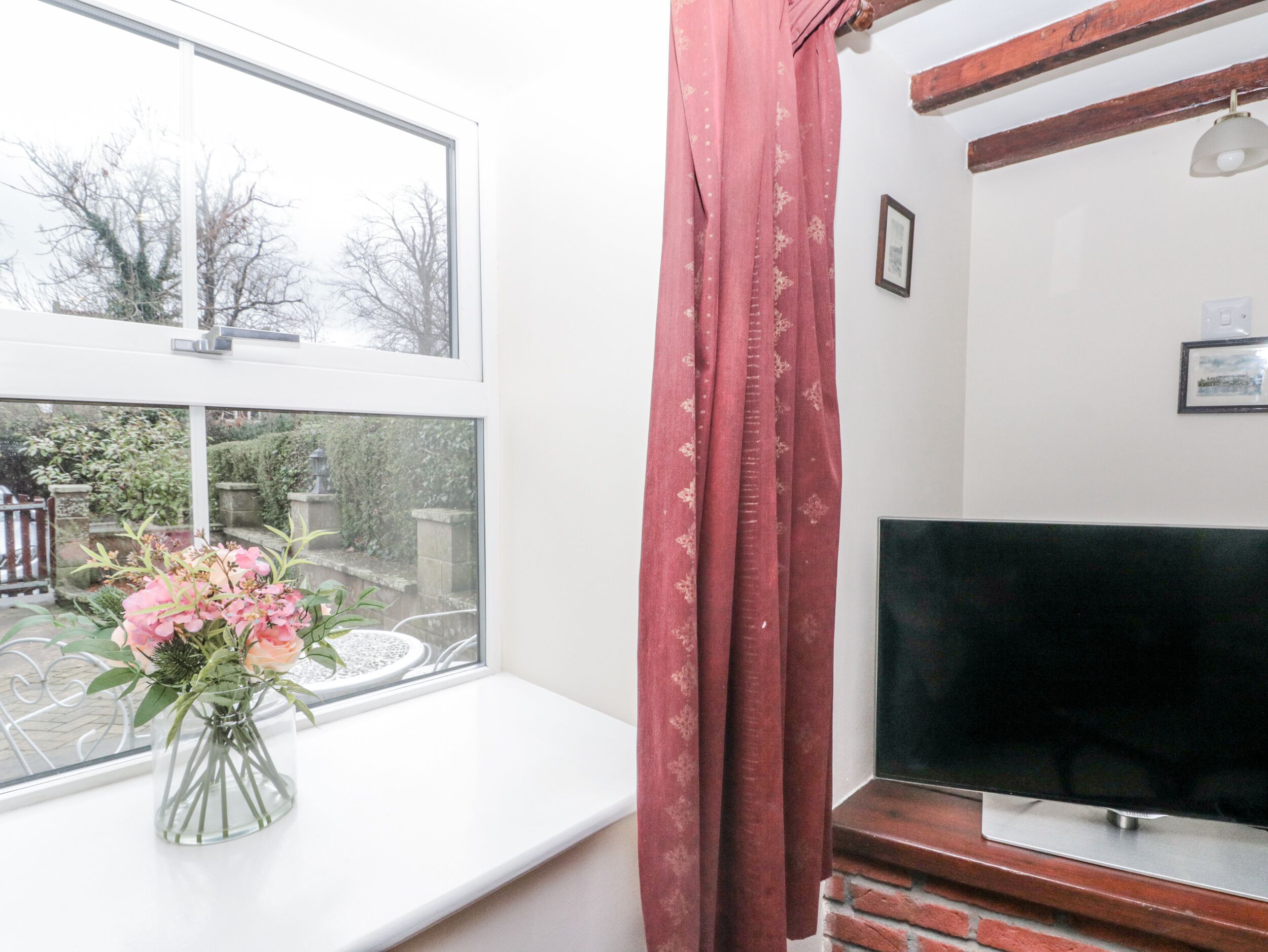A room with a TV on a wooden stand, a window with a vase of flowers, and a red curtain.