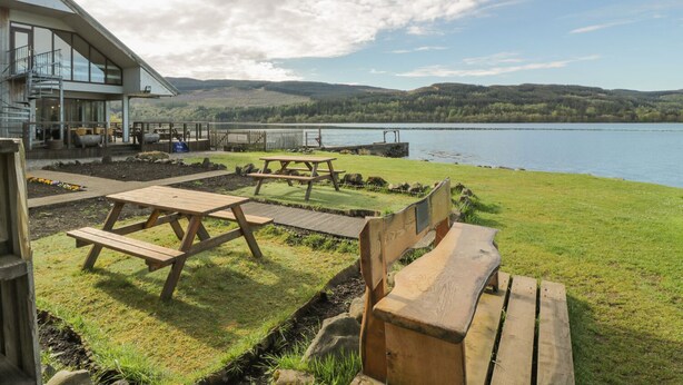 A lakeside view with picnic tables and a wooden bench, a building with large windows, and a clear sky. - Fois House (Callander)