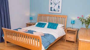A neatly made bed with a wooden frame, blue bedding, and two decorative pillows. There are two bedside tables with lamps, a wooden nightstand, and a small wooden cabinet.