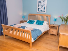 A neatly made bed with a wooden frame, blue bedding, and two decorative pillows. There are two bedside tables with lamps, a wooden nightstand, and a small wooden cabinet.