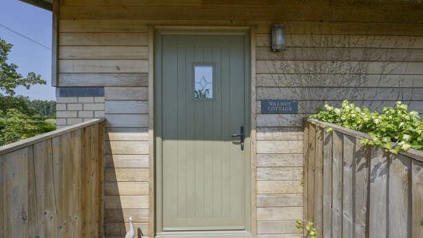 A wooden cottage with a green door, a sign reading "Walnut Cottage," and a small garden area with a wooden fence and greenery.