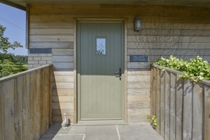 Una cabaña de madera con puerta verde, un letrero que dice 'Walnut Cottage' y un pequeño jardín con cerca de madera y vegetación.