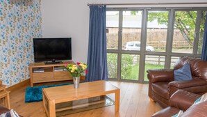 A living room with a leather sofa, a wooden coffee table, a TV on a wooden stand, and a large window with a view of a car and trees.