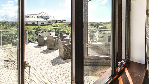 A view from a room with a large window overlooking a deck with wicker furniture and a house in the background. - The Gables (Holyhead)