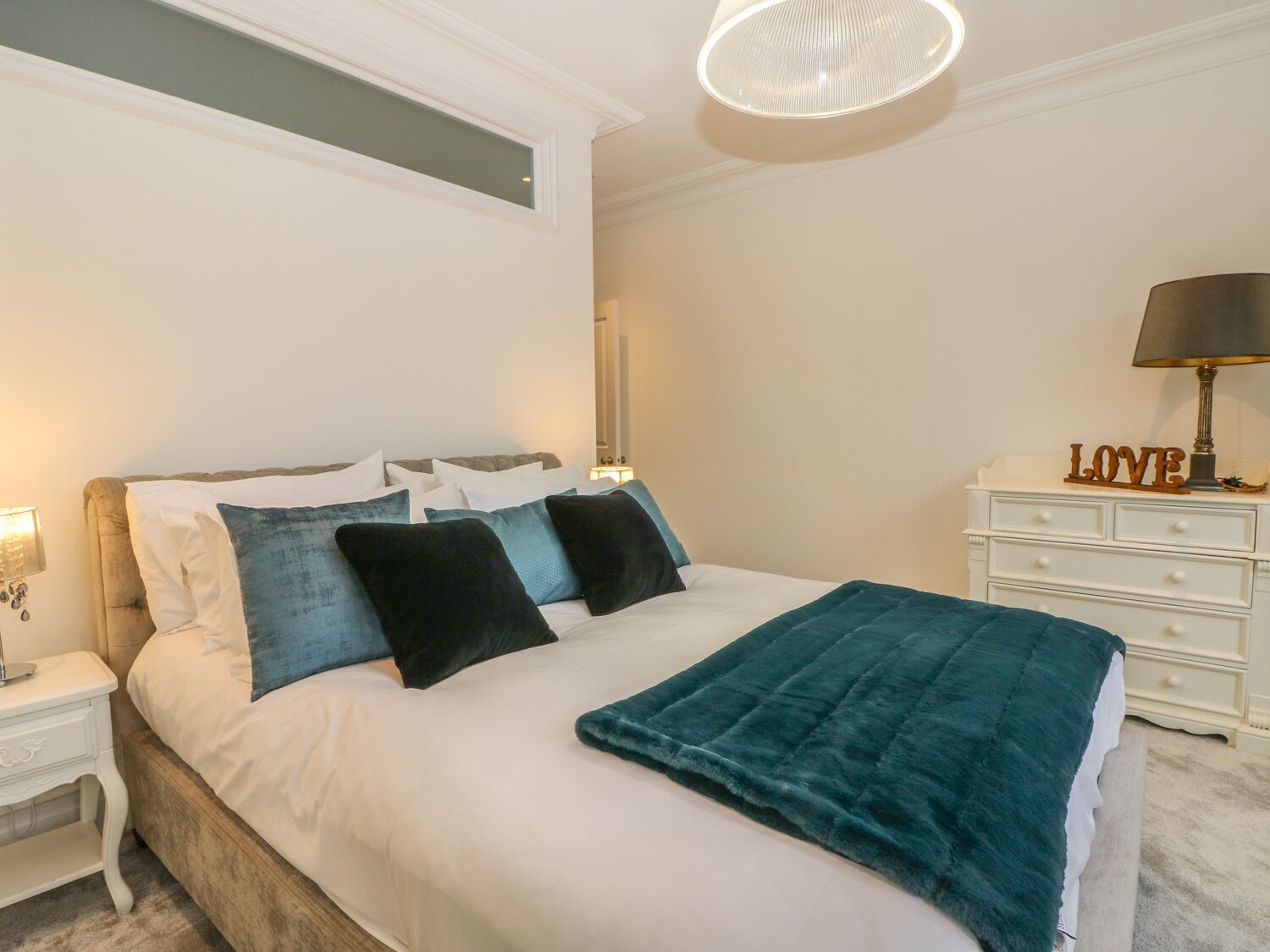 A neatly made bed with white and teal bedding, a teal throw blanket, and a bedside table with a lamp and a decorative item spelling "LOVE".