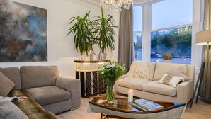 A modern living room with a grey sofa, a beige sofa, and a glass coffee table. There is a large window with a view of the outside, a chandelier, and a painting on the wall.