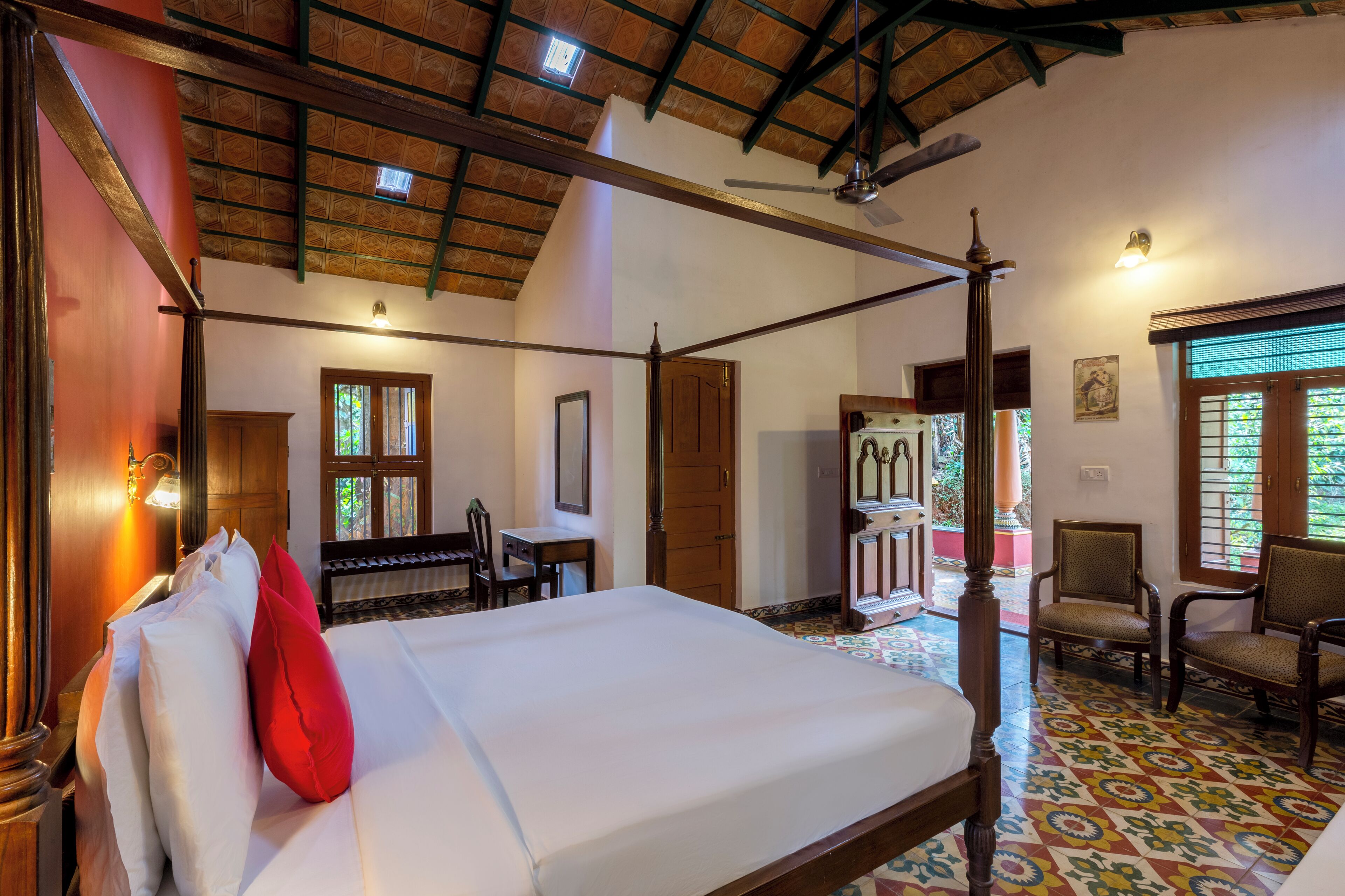 A four-poster bed with white linens and a red pillow, wooden chairs, a desk, and a ceiling fan in a room with patterned floor tiles and a sloped ceiling.