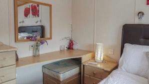 A bedroom with a bed, a bedside table with a lamp, a dresser, a mirror, and a wall with a floral painting. - The Cedars (Carnforth)
