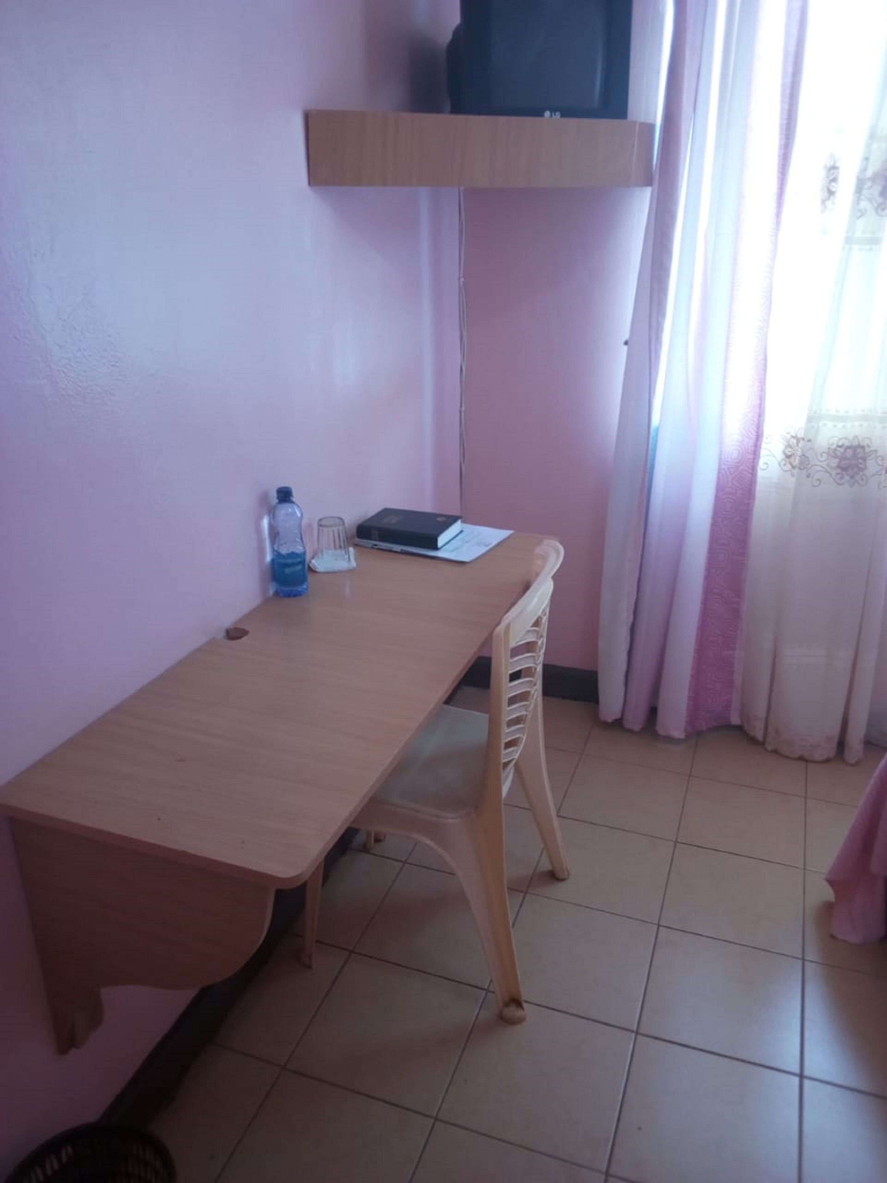 Une petite pièce avec un bureau en bois, une chaise, une bouteille d’eau, un livre et un téléviseur posé sur une étagère.