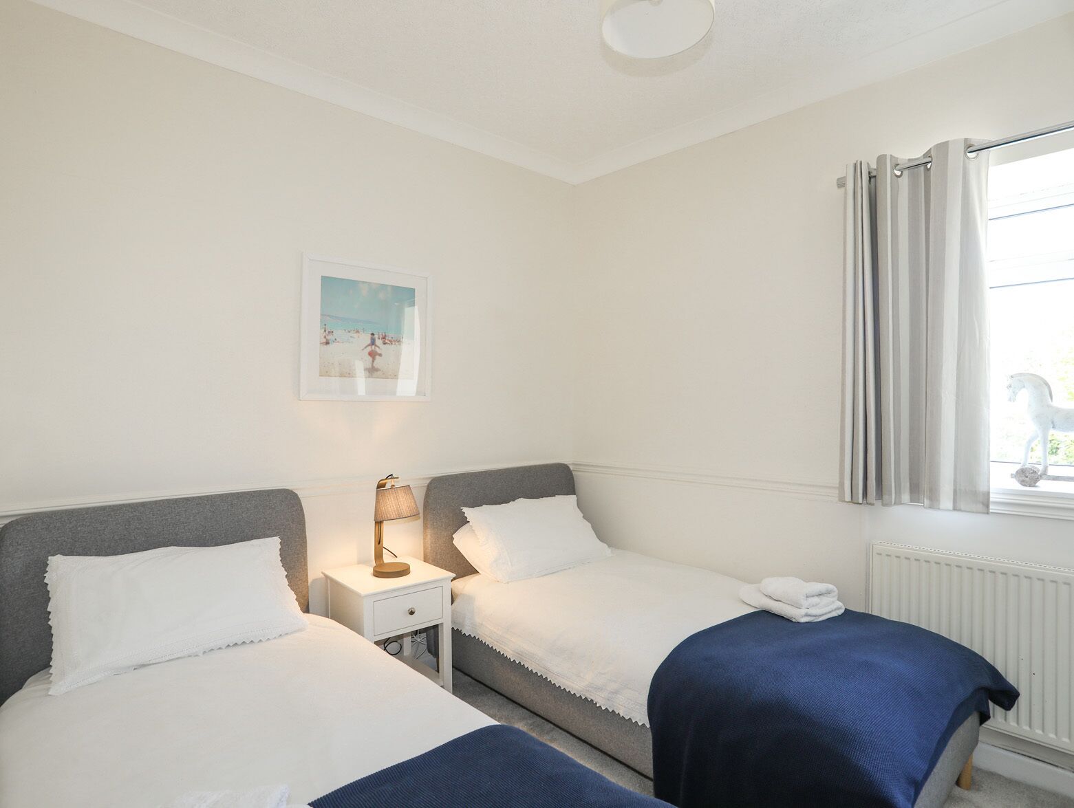 A hotel room with two single beds, a nightstand with a lamp, a framed picture on the wall, and a window with curtains.