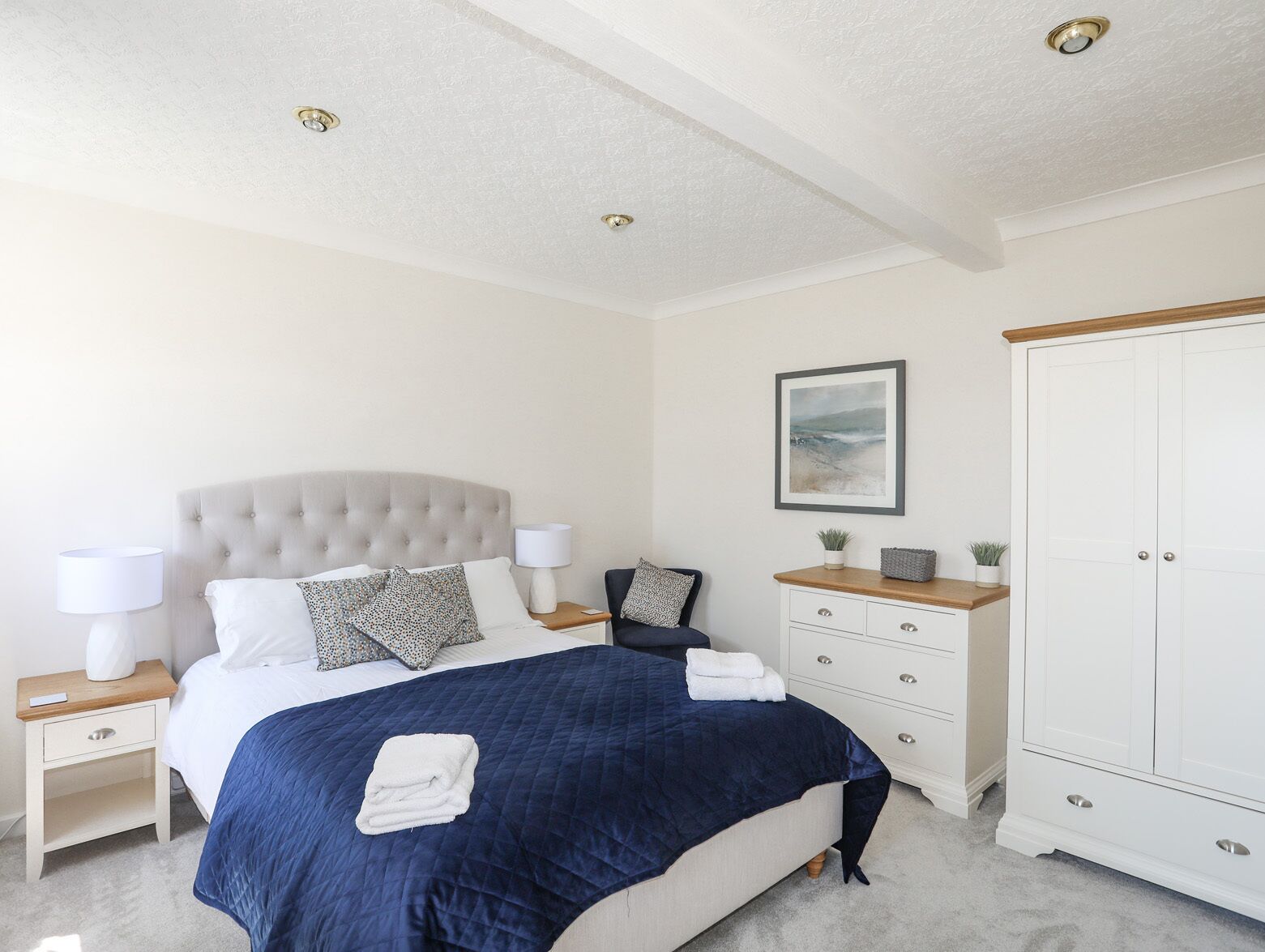 Une chambre à coucher avec une tête de lit capitonnée, un grand lit recouvert d’une courtepointe bleue, deux tables de chevet avec des lampes, une commode, une armoire et un tableau encadré au mur.