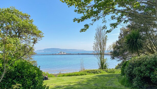 A grassy area with a view of a calm body of water and distant hills. - Sea Pebbles (Weymouth)