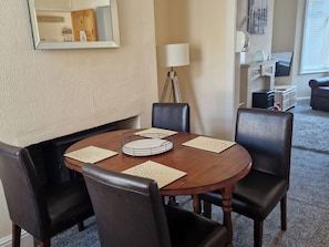 Uma sala de jantar com uma mesa de madeira redonda, quatro cadeiras pretas, um abajur e uma televisão ao fundo.
