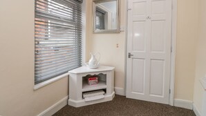 Una habitación de esquina con una puerta blanca, una ventana con persianas, una pequeña mesa consola blanca con una jarra y una cesta, y un espejo en la pared.